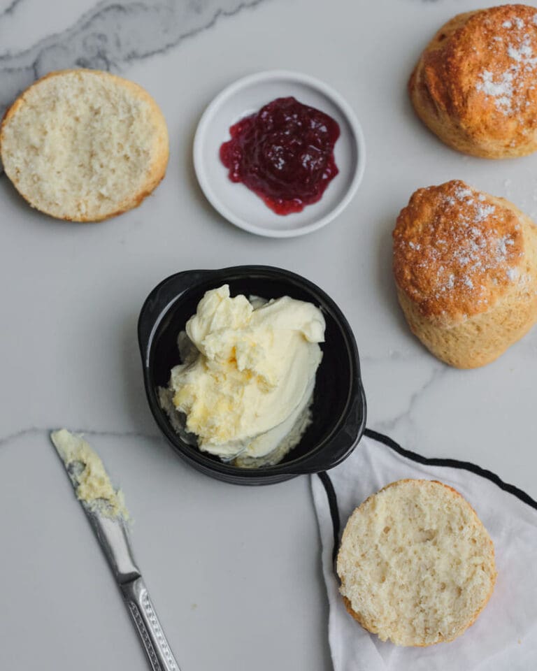 An overhead view of freshly baked scones, some halved and ready to be topped, accompanied by a dish of clotted cream, a small plate of red jam, and a butter knife. This simple, elegant flat lay captures the essence of national cream tea day, celebrating the beloved British tradition in style.